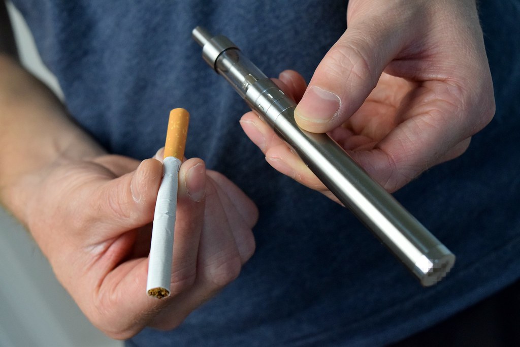 Hand holding a vape pen beside a cigarette